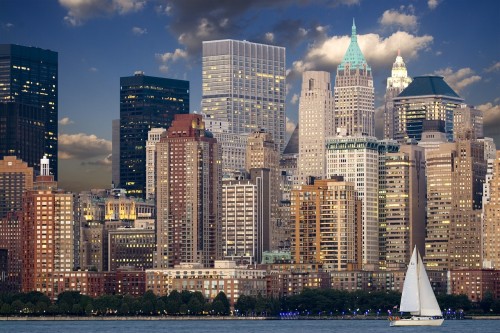 A sailboat sails in NYC's Hudson River in front of Manhattan skyscrapers.