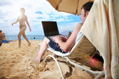 A technomad AKA a digital nomad is working remotely on a laptop while sitting on a beach chair in the sand of on an exotic island.