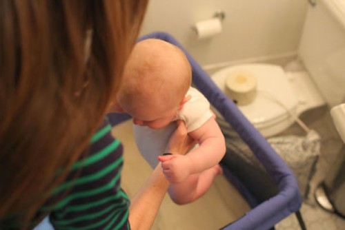 A mother is picking up her baby from a crib that's in a bathroom.