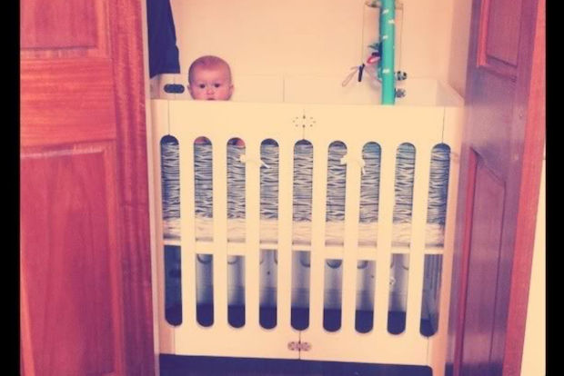 A baby is peeking over the top of a crib that's stored in a closet.