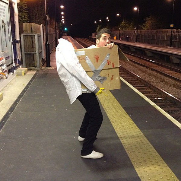 A man is wearing a head-in-box Halloween costume and standing on a platform at a train station.