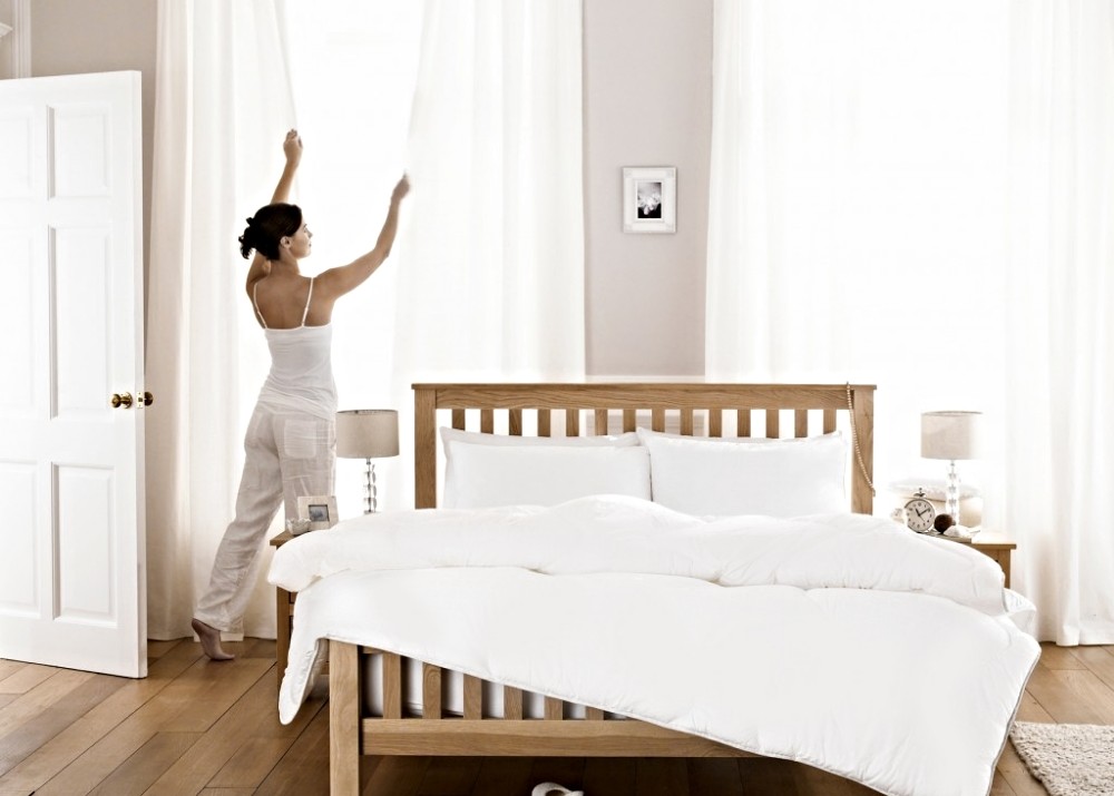 A woman dressed in white is closing her white bedroom curtains.