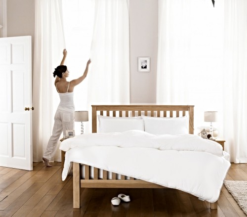 A woman dressed in white is closing her white bedroom curtains.