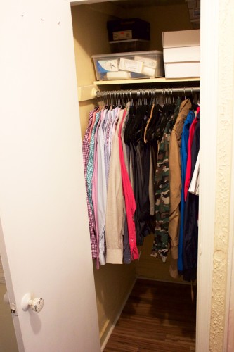 An organized, decluttered bedroom closet with shirts, jackets, and pants hanging and storage containers stacked neatly on a shelf.