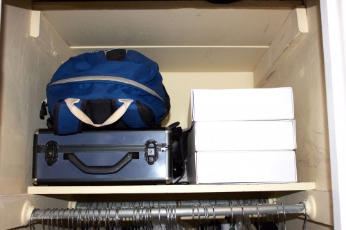 A blue backpack, a blue storage container, and three white boxes stacked on an unorganized bedroom closet shelf.