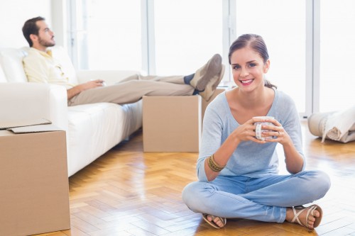 A woman is sitting on the floor of an apartment and holding a coffee mug while a man is sitting on a couch with his feet atop moving boxes.
