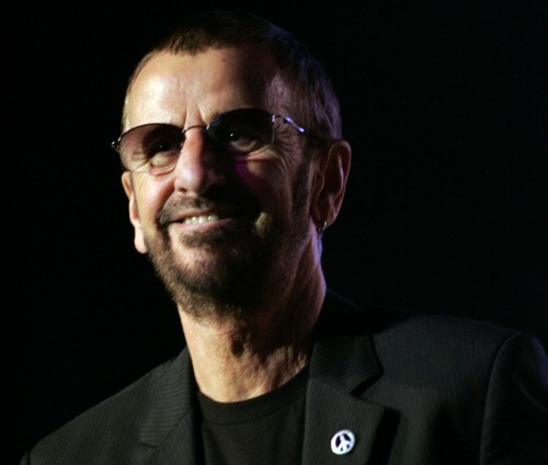The Beatles drummer Ringo Starr is holding a microphone and wearing sunglasses, a blazer, and a t-shirt.
