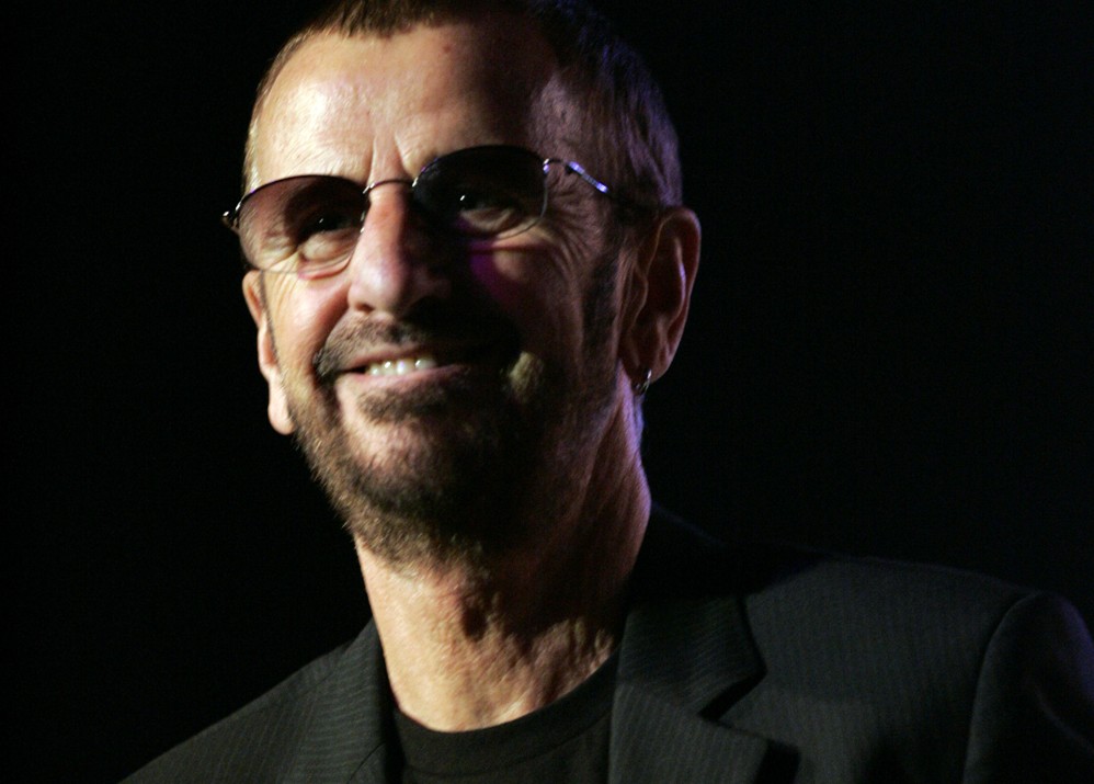 The Beatles drummer Ringo Starr is holding a microphone and wearing sunglasses, a blazer, and a t-shirt.