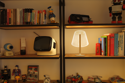 Studio Cheha's 2D/3D CLASSi BULBING lamp is on a storage shelf next to books, a retro TV, an old video game console, Batman, Joker, Donald Duck, robots, a red telephone box, and other toys.