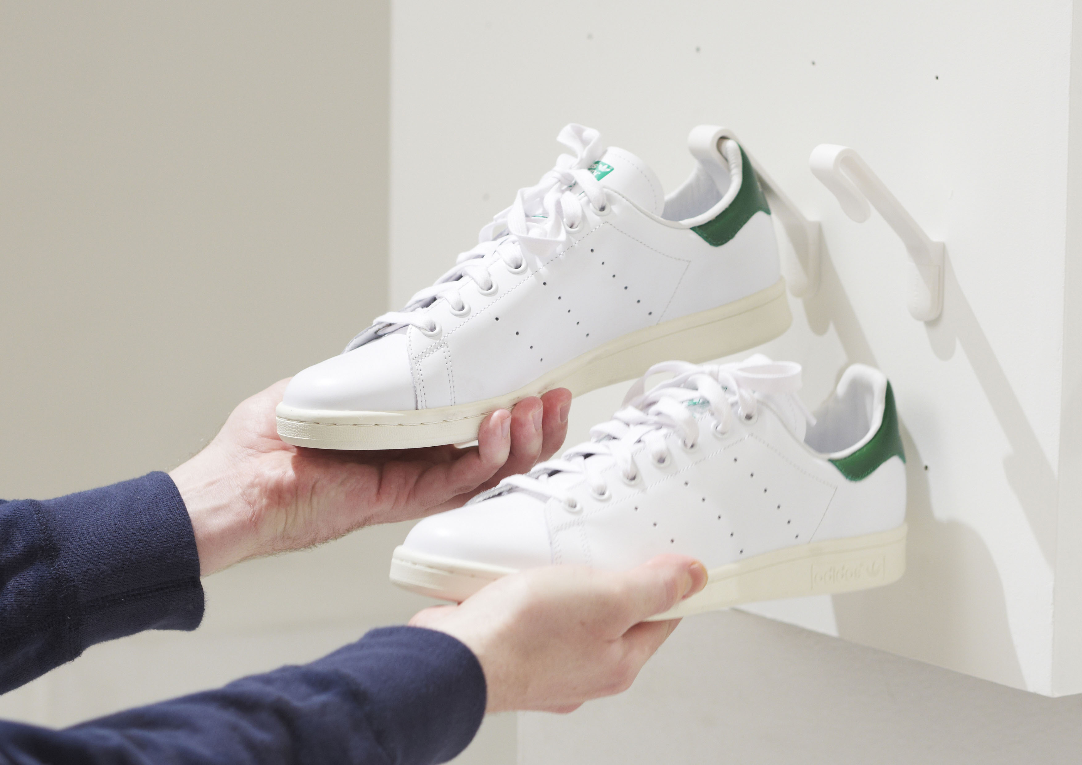 Two hands are taking a pair of white Adidas Stan Smith sneakers off of two white Staekler shoe storage hooks.
