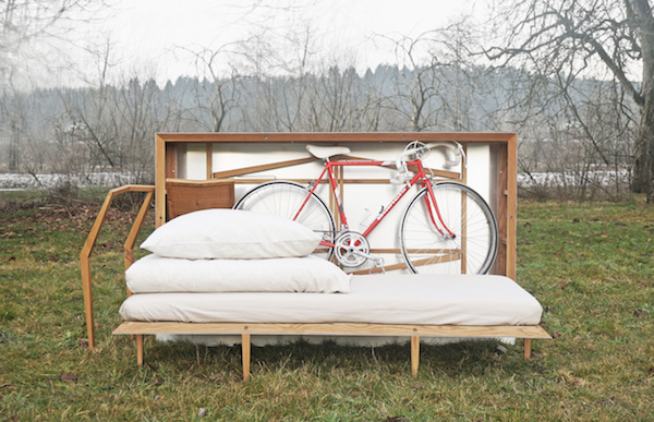 JUUST Design's Travelbox is standing in the grass and open, showing a mattress, table, chair, bike, and pillows in storage.