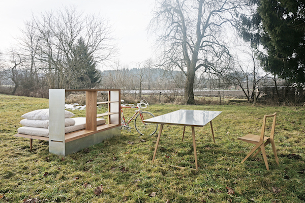 An open Travelbox with a bed, pillows, and storage shelves is standing outside in the grass and surrounded by a bike, table, chair, and trees.