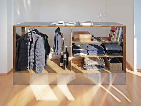 An open Travelbox is inside an apartment and storing jackets, pants, shirts, sweaters, wine bottles, and books on storage shelves.