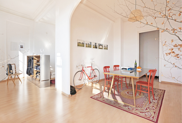 Travelbox is inside of a clean, minimally-decorated tiny apartment, storing clothes, and surrounded by a chair, bike, dining table, and chairs.