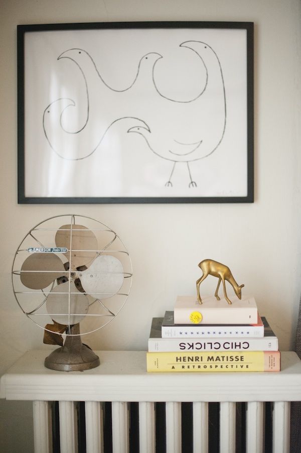 A white radiator shelf is storing an old metal fan, four stacked books, and a brass deer figurine on top.