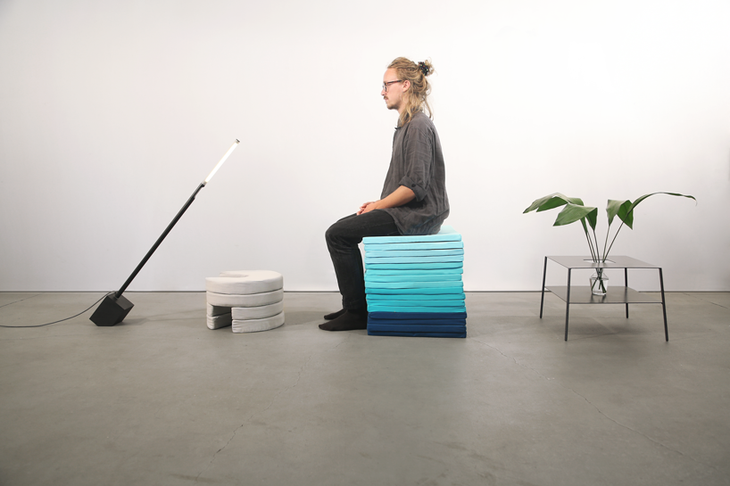 A man is sitting on stacked square Pile cushions and surrounded by a floor lamp, steel coffee table, and a footrest made of stacked circular Pile cushions.