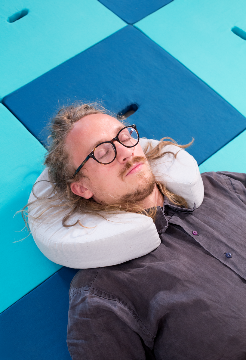 A man with glasses and long blonde hair is wearing a circular Pile cushion as a neck pillow and lying on a bed made of square Pile pieces. 