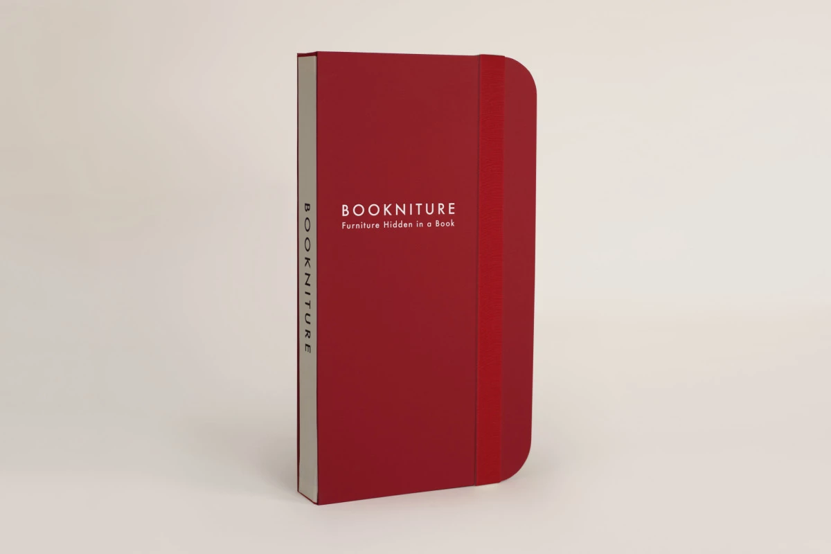 A closed red Bookniture, which looks like a book and contains space-saving furniture, is standing on a white surface.