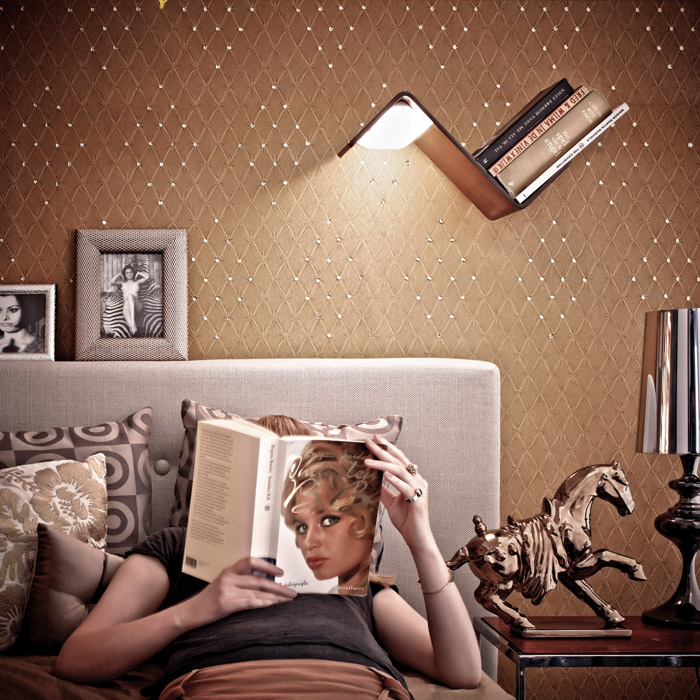 A LiliLite, which is a wooden bookshelf, bookmark, and lamp that mounts to a wall, is shining over a woman reading a book in bed.