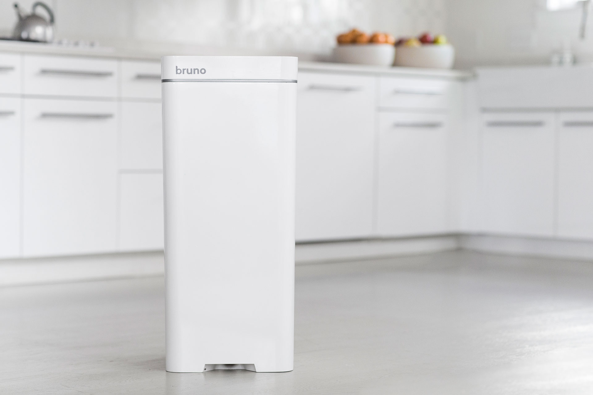 A white BrunoSMART trashcan/vacuum is inside of a white kitchen in a tiny apartment.