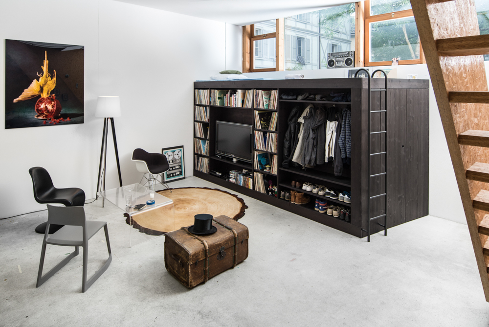 A black multi-functional Living Cube, which is a media center, bar, closet, bike rack, bed, bookcase, desk, and storage unit, is in the corner of a modern living room.