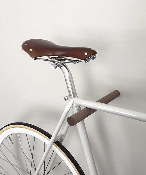 A brown fluo wooden bike hook is mounted to a white wall and hanging a white bicycle with a brown leather seat.