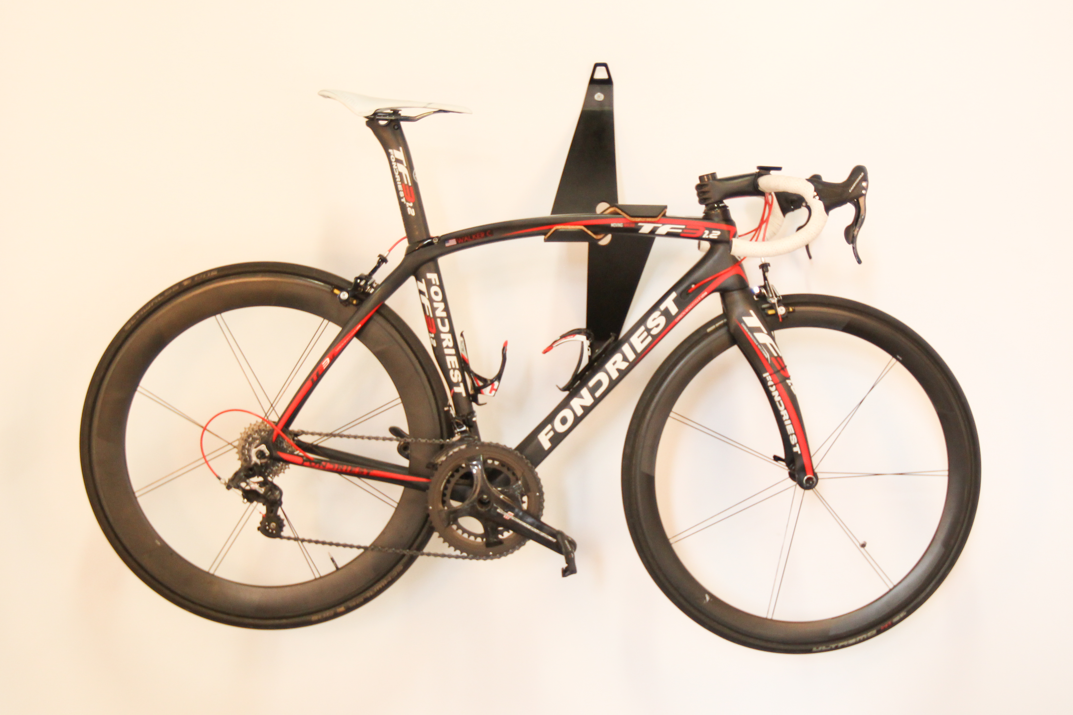 A black Reclamation Art and Furniture Bike Valet, which looks like airplane wings, is mounted to a white wall and hanging a red and black bicycle.