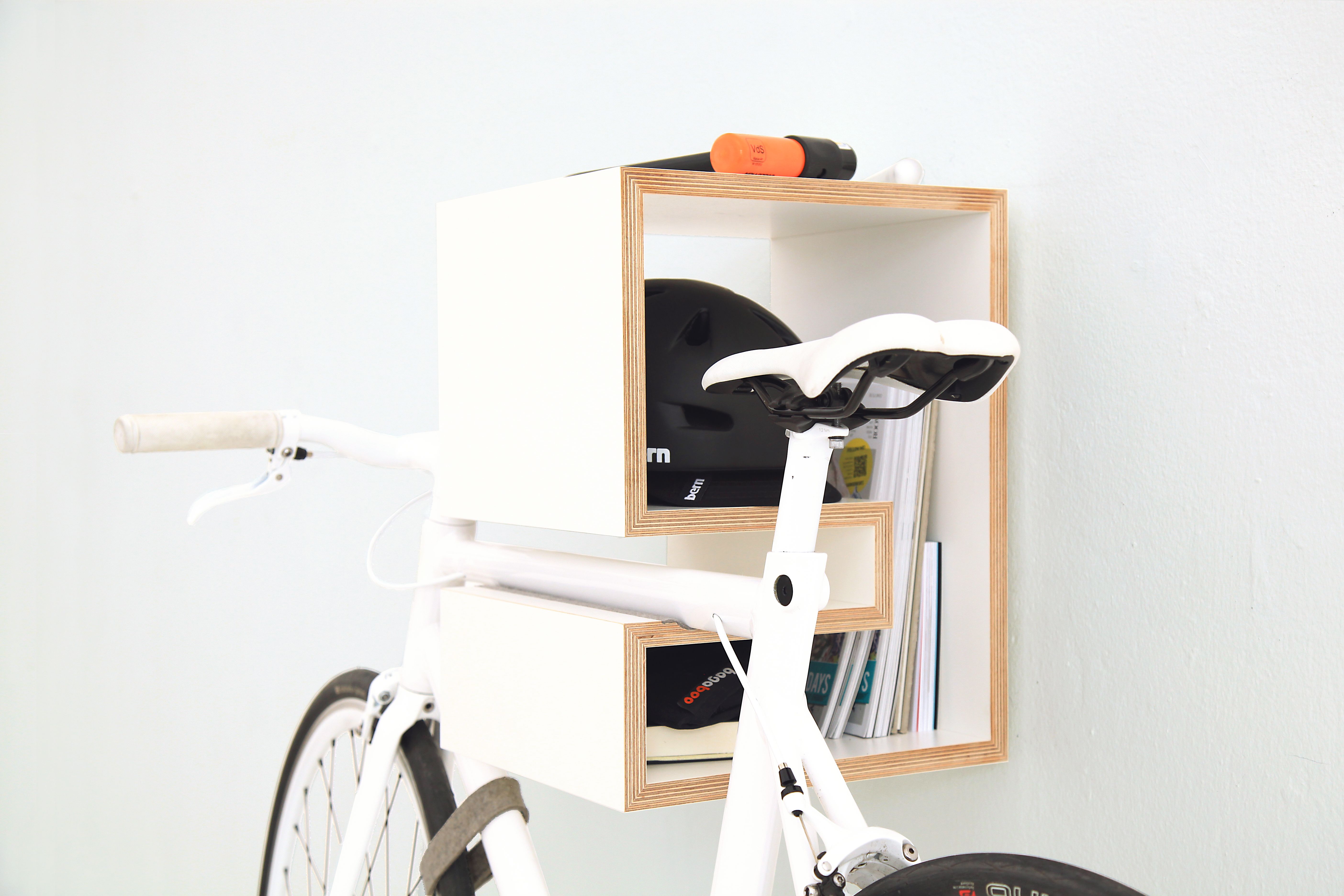 A white Mikili Kappo bike storage rack is storing a white bicycle, black bike helment, black U-Lock, and various books.