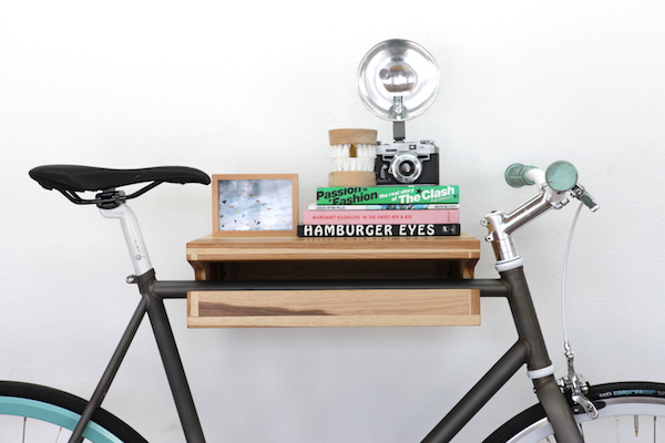 A Knife & Saw white oak Bike Shelf is mounted to a white wall and storing a black bike, books, a framed picture, fake teeth, and a vintage camera.