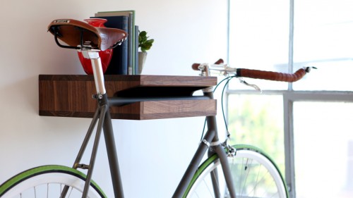 A walnut Knife & Saw Bike Shelf is storing a bicycle, red vase, four books, and a small plant.