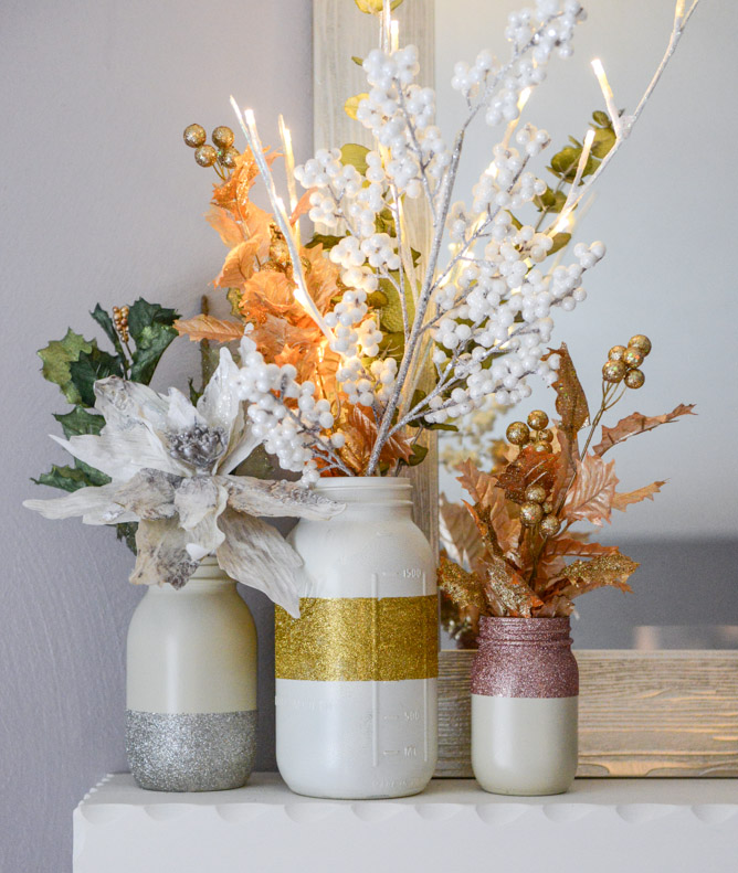 A DIY decoration made of 3 white glittery Mason jars and various artificial flowers and branches with leaves.