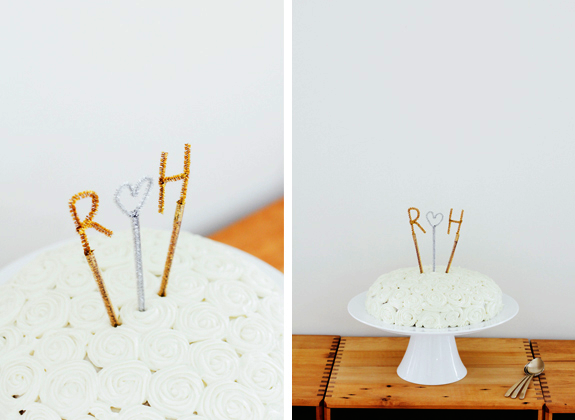 A DIY decoration of cake toppers made from pipe cleaners are popping out of the top of a white-frosted cake on a white cake stand on a wooden surface.