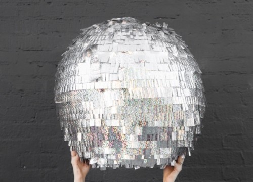 2 hands are holding up a DIY NYE disco ball pinata in front of a dark gray brick wall.