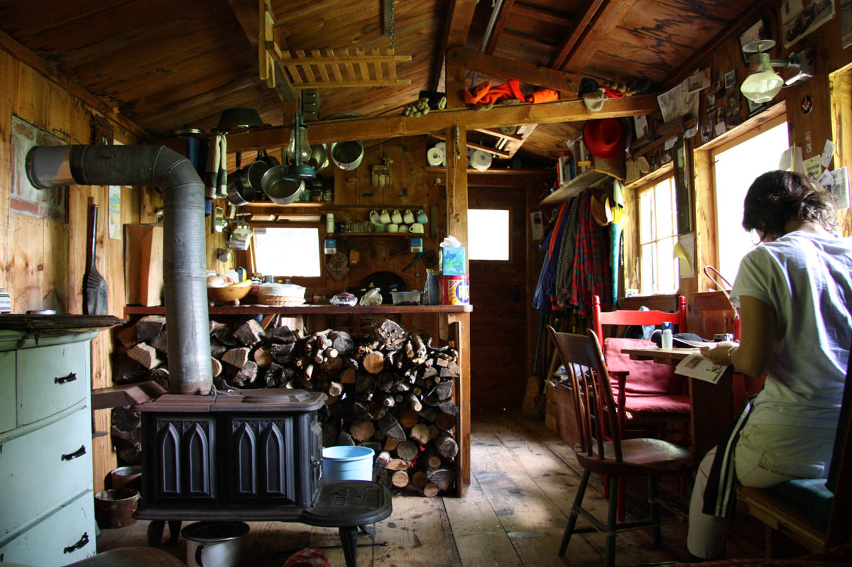 A woman is reading at a table in a minimally decorated cabin with a wood burner stove.
