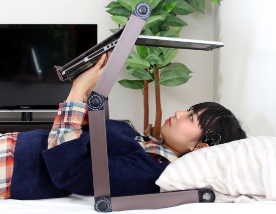 A woman is laying in bed and typing on a laptop attached to a bed-desk.