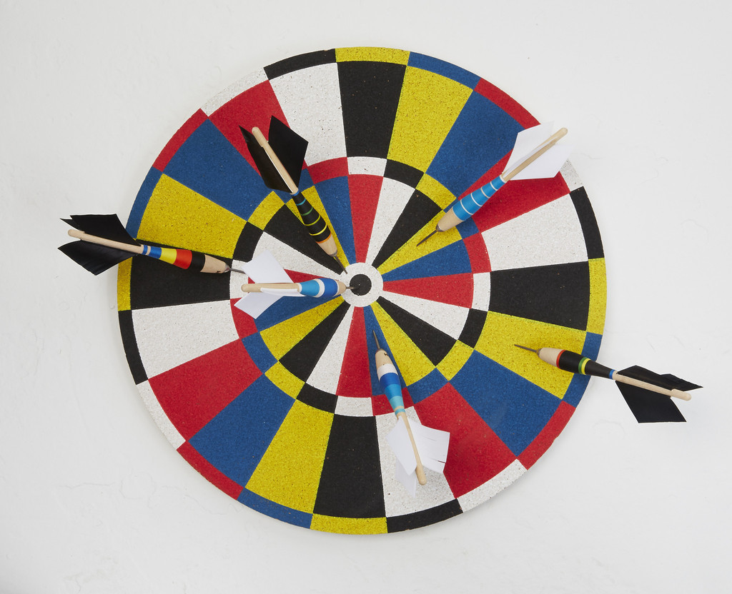 A Fredericks & May dartboard mounted to the wall in a small apartment with multiple darts poking into it.