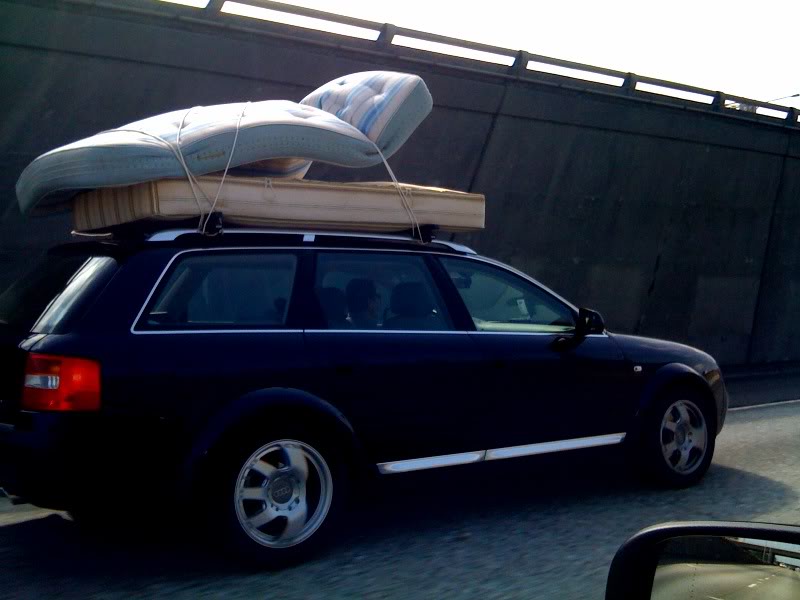 2 mattresses tied to the top of an Audi station wagon driving on the highway.