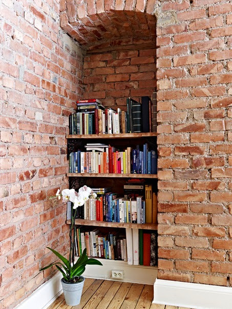 A DIY bookcase in a tiny apartment's exposed brick nook is a prime example of a cool book storage hack.