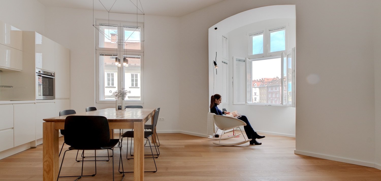 A lady is reading a book while sitting in a rocking chair in front of a window in a clean apartment.