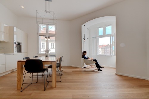 A lady is reading a book while sitting in a rocking chair in front of a window in a clean apartment.