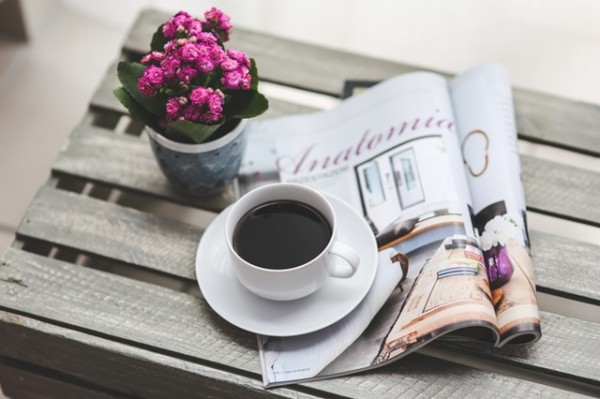A magazine, a flower in a pot, and a cup of coffee are on top of a wooden crate.