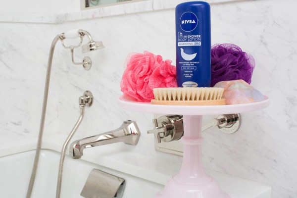 cake stand on top of a bathtub and storing loofahs, nivea in-shower body lotion, a scrubber, and soap