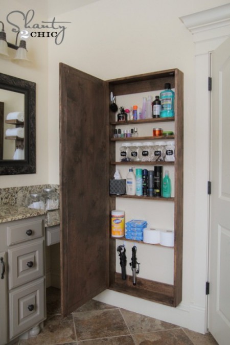 a wooden diy bathroom storage cabinet is storing listerine, rubbing alcohol, nail polish, clorox wipes, curling irons, aveeno lotion, and more
