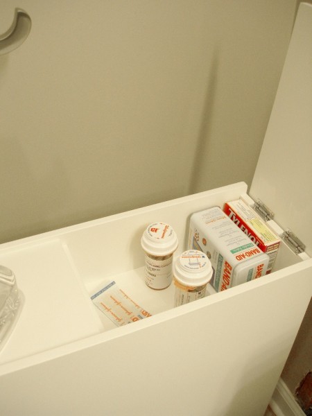 free-standing cabinet's top hidden storage space is storing walgreens prescription bottles, a bandaid can, wrapped bandaids, and a tylenol box