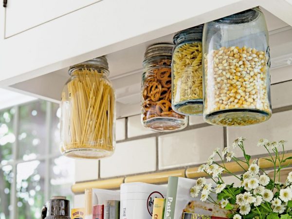 4 mason jars screwed under a kitchen cabinet are storing pasta, pretzels, and popcorn kernels 
