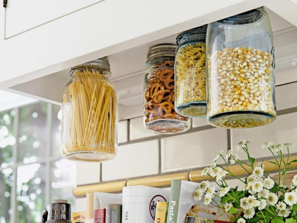 4 mason jars screwed under a kitchen cabinet are storing pasta, pretzels, and popcorn kernels