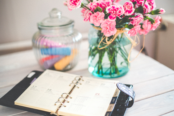 a personal organizer, pink flowers in a glass vase with water, and a glass jar containing chalk are on top of a wooden desk