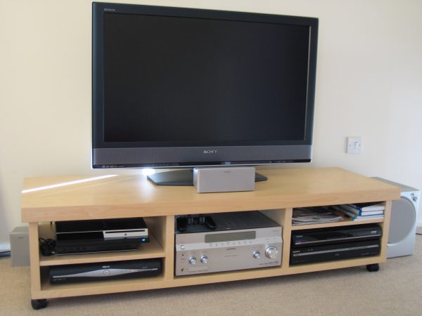 a sony flat screen tv stand with wheels is storing a sound system, speaker, playstation console, playstation controller, cable box, books, and papers
