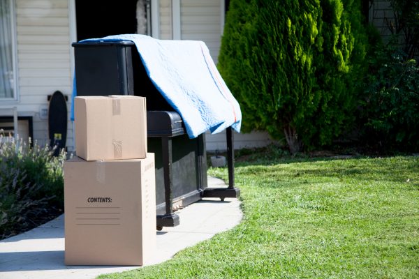 piano, moving blanket, and storage boxes on a walkway in front of a house on a sunny day
