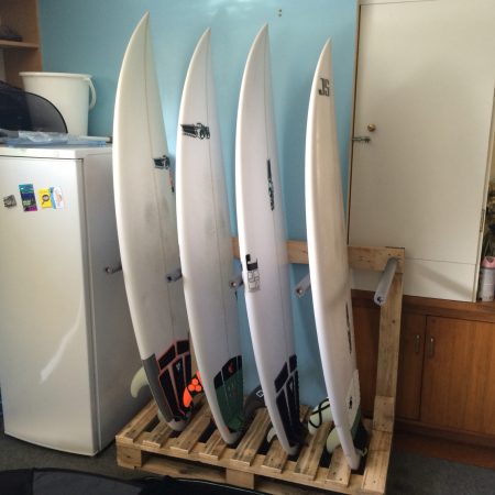 A DIY pallet surfboard rack storing 4 white surfboards vertically in a kitchen next to a fridge